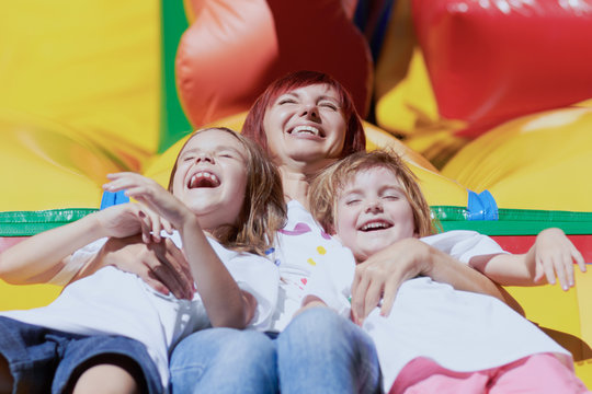 Mother And Her Daughters Having Fun On Jumping Castle At Playground In Sunny Summer Day.Happy Family Together.Two Little Girls Laughing Out Loud With Their Beloved Mommy On Inflatable Bouncing Castle