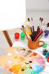 Wooden easel with clean paper and art supplies in room