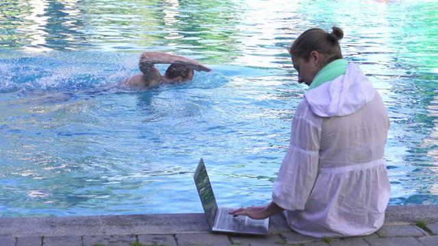 Woman with laptop sit by the pool, man swimming