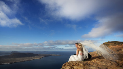 Bride and groom sitting on ocean shore, volcanic landscape.