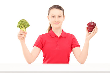 Smiling female teenager sitting and holding apple and broccoli
