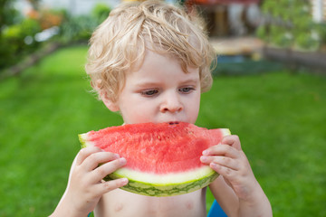 Adorable little toddler boy with blond hairs eating watermelon i