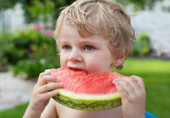 Adorable little toddler boy with blond hairs eating watermelon i