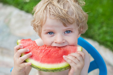 Adorable little toddler boy with blond hairs eating watermelon i