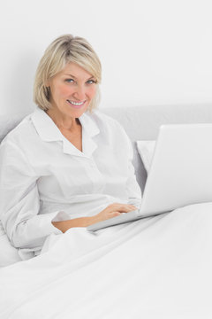 Blonde woman sitting in bed with laptop smiling at camera