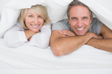 Cheerful couple smiling under the covers at the camera