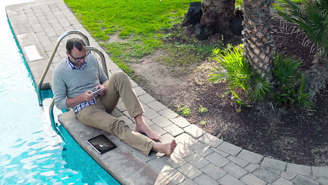 Young man with cellphone and tablet sitting by swimming-pool, vi