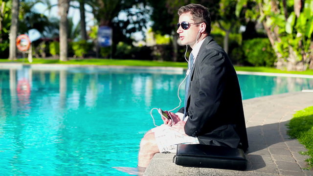 Businessman sitting by the swimming pool and listening music 