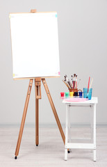Wooden easel with clean paper and art supplies in room - obrazy, fototapety, plakaty