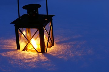 Lantern with burning candle on snow in the evening.