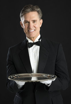 Confident Waiter In Tuxedo With Tray Standing Against Black Back