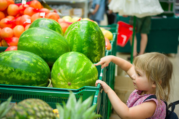Fototapeta premium Adorable girl with backpack select watermelon in supermarket