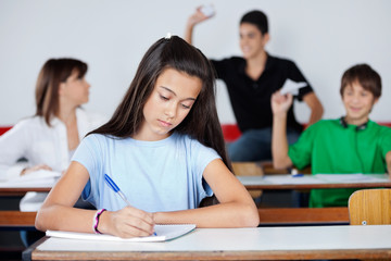 Schoolgirl Writing At Desk While Classmates Playing