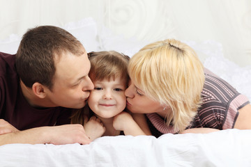 Happy father and mother lie on white double bed and kiss little