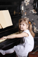 Little beautiful girl in white dress sits at black piano with mu