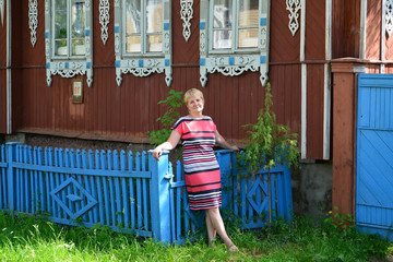 The woman stands near the wooden house with carved platbands