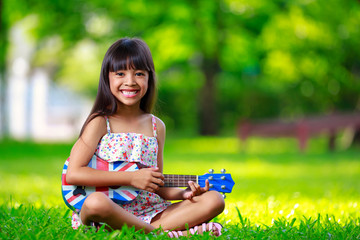 Little asian girl sitting on grass and play ukulele