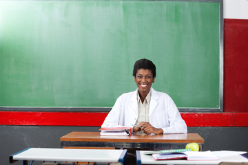 Happy Teacher Sitting At Desk In Classroom