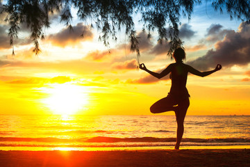 Silhouette young woman practicing yoga on the beach at sunset.