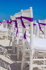 Fototapeta na wymiar White wedding chairs decorated with purple bows on white sandy