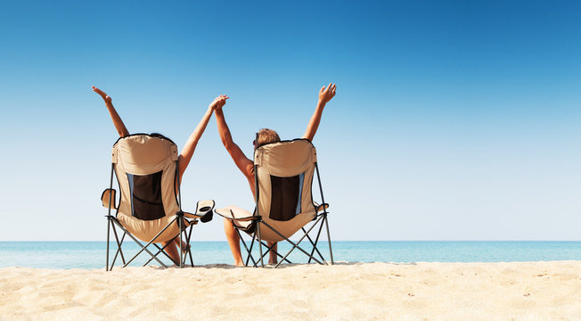 Young Couple Sitting In Cozy Chairs On The Wild Sea Beach And Re