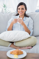Obraz na płótnie Canvas Cheerful woman on sofa drinking coffee and having croissant