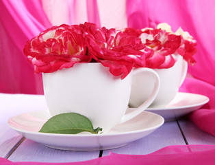 Roses in cups on wooden table on pink cloth background