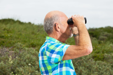Senior man with beard and glasses using binoculars outdoors in g