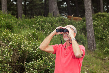 Senior retired man with beard using binoculars outdoors. Wearing