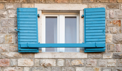 Wooden window with blue open jalousies in old gray stone wall