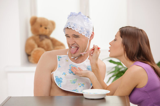 Big Baby Eating. Young Woman Feeding An Infant Adult Man In Baby