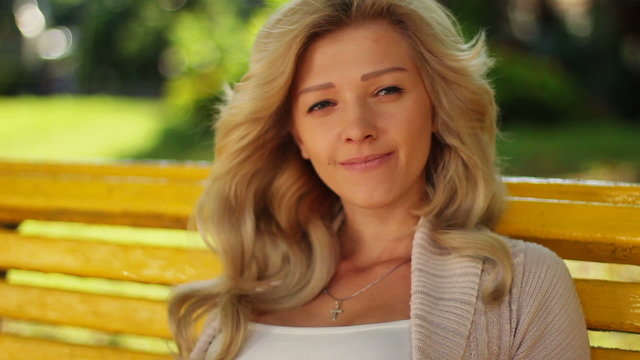Pretty blond female smiles in park on bench, looks