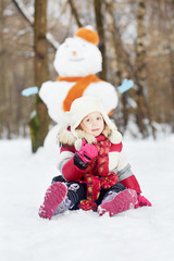 Fototapeta na wymiar Little girl sits on snow in front of big snowman in winter park,