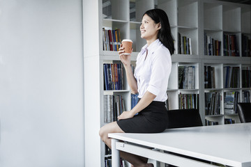 Fototapeta na wymiar Smiling young businesswoman holding a coffee and sitting on a table, looking away