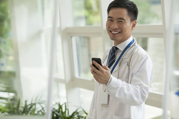 Fototapeta premium Smiling doctor using his phone in the hospital lobby, looking at camera, glass doors