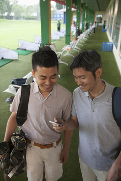 Two male friends smiling and getting ready to leave the golf course, looking down at phone