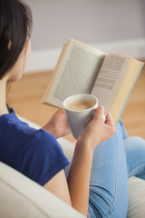Obraz premium Young woman sitting on the sofa reading a book holding her coffe