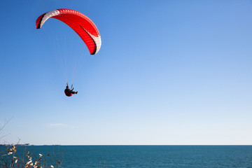 Red paraglider flying in blue sky over the ocean.