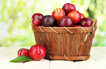 Ripe plums in basket on wooden table on natural background