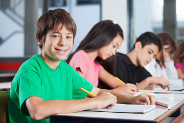 Boy Sitting At Desk With Friends Writing In Classroom