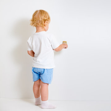 Baby boy with paint brush rear view standing near blank white wa
