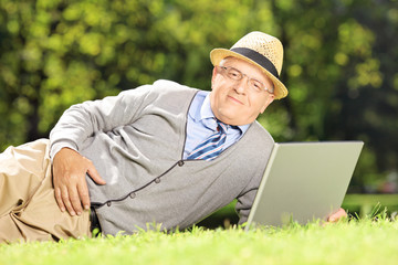 Fototapeta premium Senior man with hat lying on a grass working on a laptop in park
