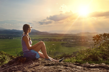 Naklejka na ściany i meble Girl sits on edge of cliff looking at sun and mountains