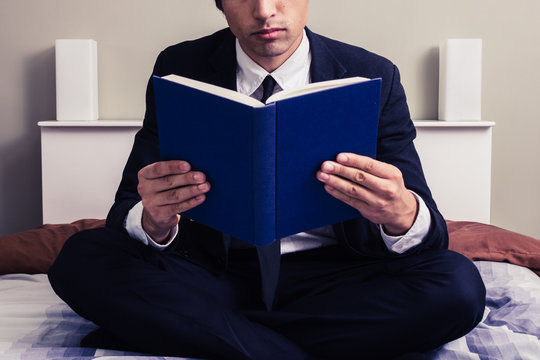 Serious young businessman sitting in bed reading book