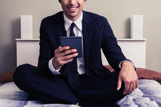 Happy young businessman sitting in bed reading on tablet