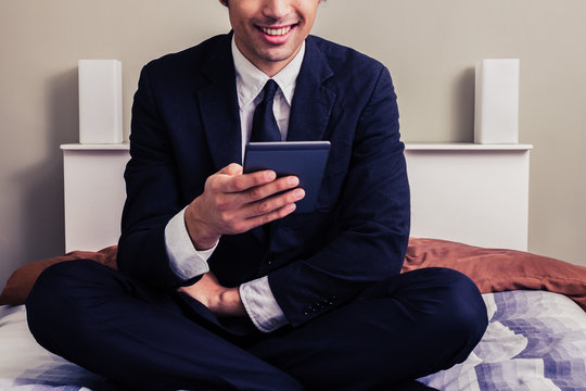 Happy young businessman sitting in bed reading on tablet