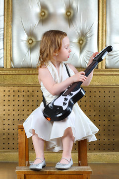 A small girl in white dress sitting in a chair with guitar