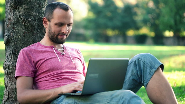 Young man with modern laptop by the tree in the park