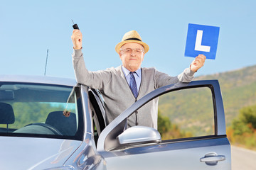 Smiling senior man posing next to his car holding a L sign