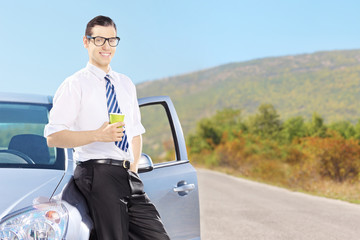 Smiling young man on his automobile drinking coffee on a road